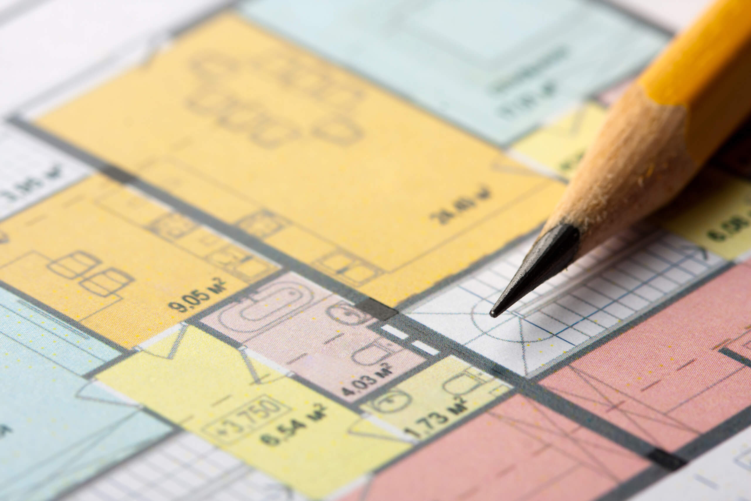 Close-up of a floor plan with various colored sections indicating different rooms and measurements in square meters. A sharpened pencil is placed on the right side of the image, pointing towards the plan as if ready for marking or note-taking, perhaps planning where to install a TV wall mounting.