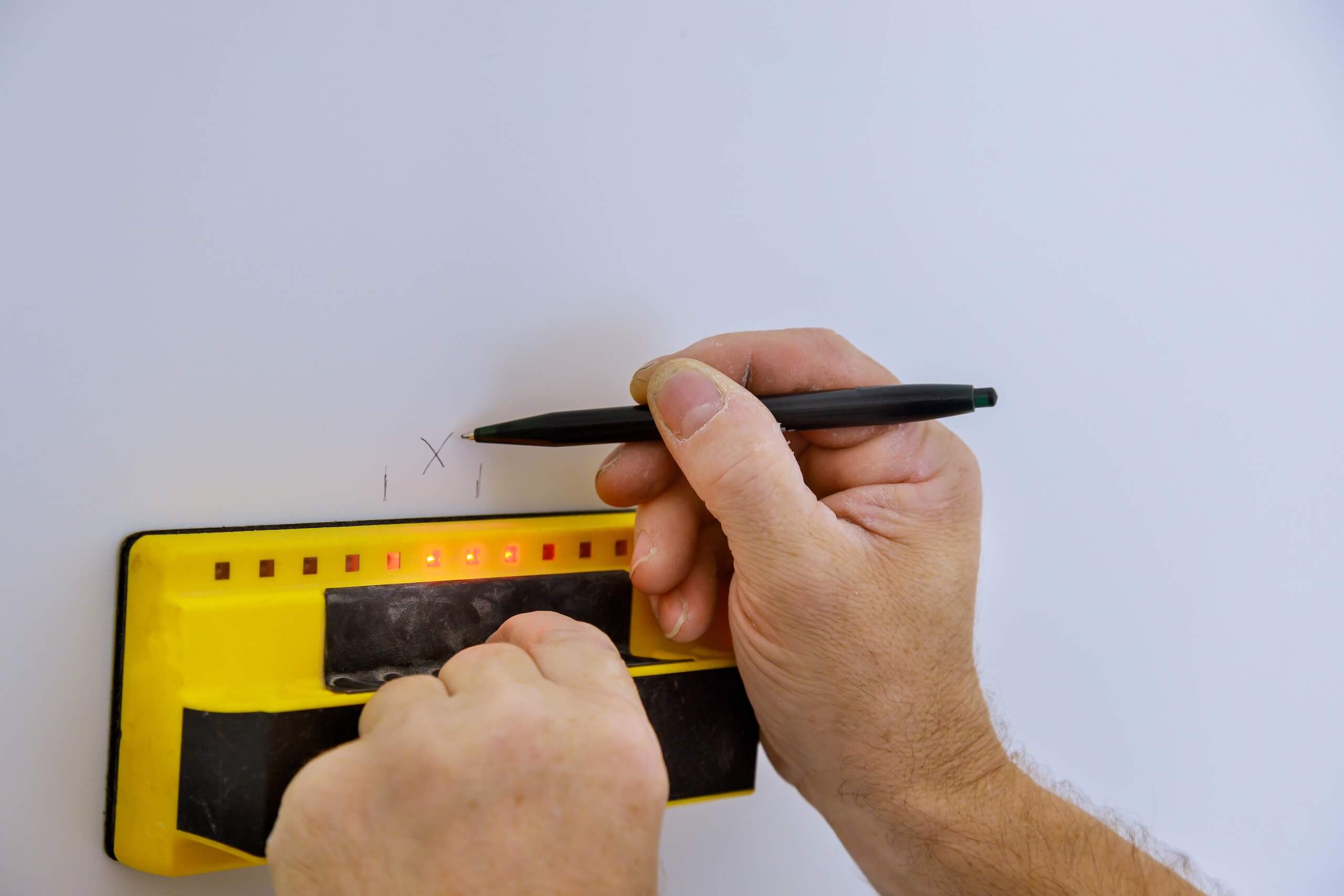A person is using a yellow stud finder on a white wall, preparing for TV wall mounting. The stud finder's red indicator lights are illuminated as the person marks the precise spot with a black pencil or pen.