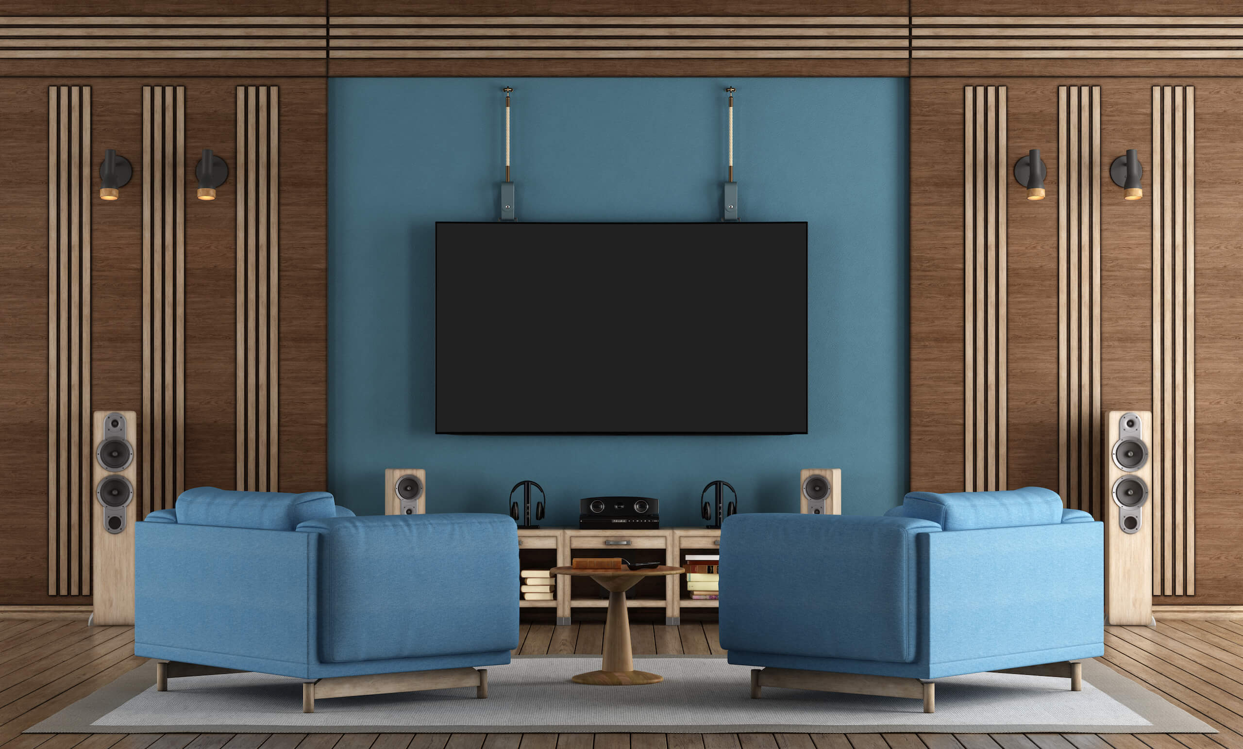 A modern home theater setup with two blue armchairs facing a large flat-screen TV mounted on a blue accent wall. The room features wooden paneling, wall-mounted speakers, and a small wooden table between the chairs. Additional speakers and audio equipment sit below the securely TV wall mounting system.
