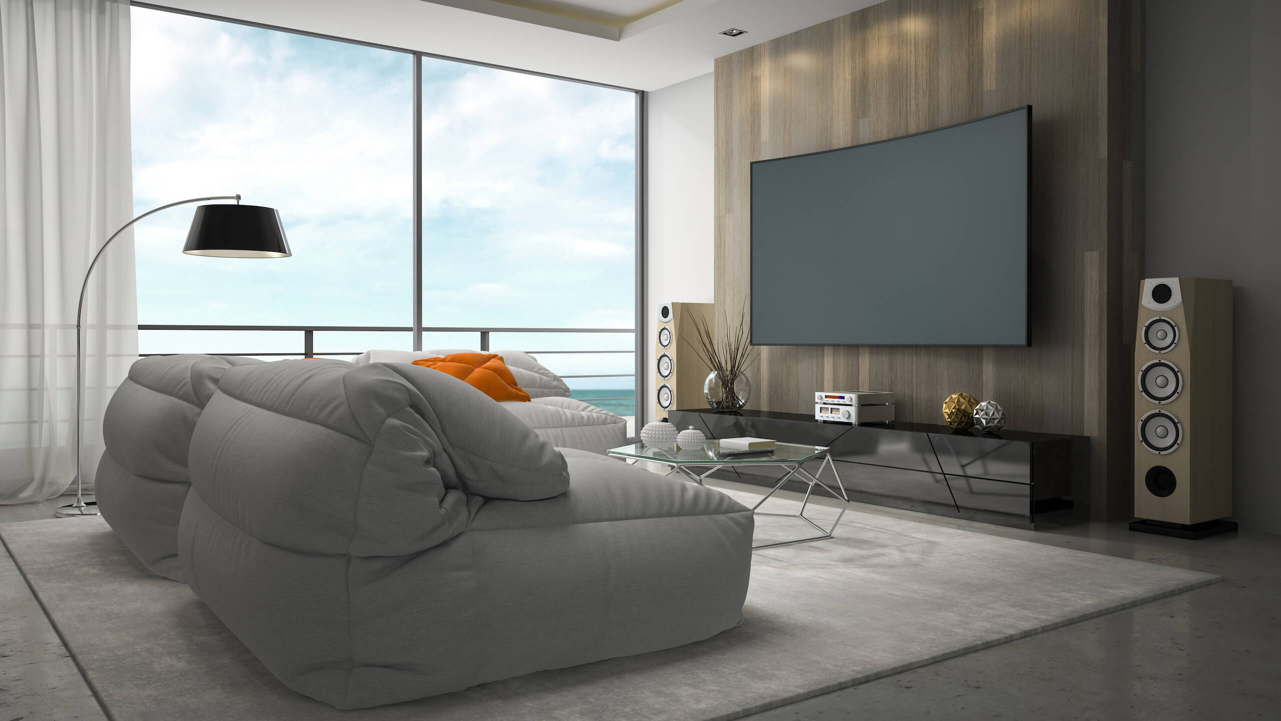Modern living room with large floor-to-ceiling windows showcasing an outdoor view. Features include a curved flat-screen TV with TV wall mounting on a wooden accent wall, two cushioned gray sofas, a black floor lamp, orange throw pillows, and a glass coffee table on a light rug.