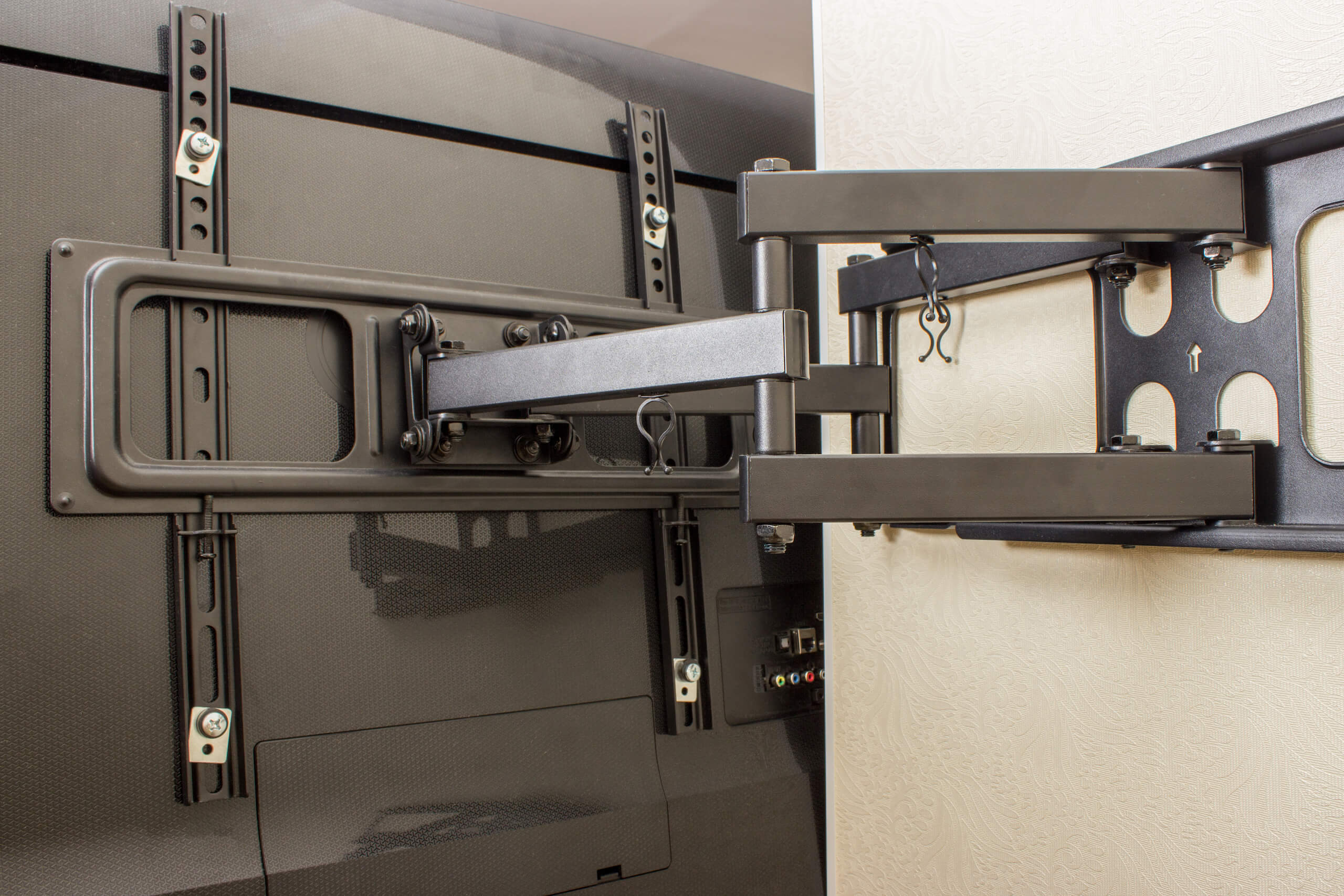 A flat-screen TV is mounted onto a wall using an adjustable, black articulated wall mount bracket. This TV wall mounting setup allows for tilting and extending the TV away from the wall. The TV's backside and the wall are visible.