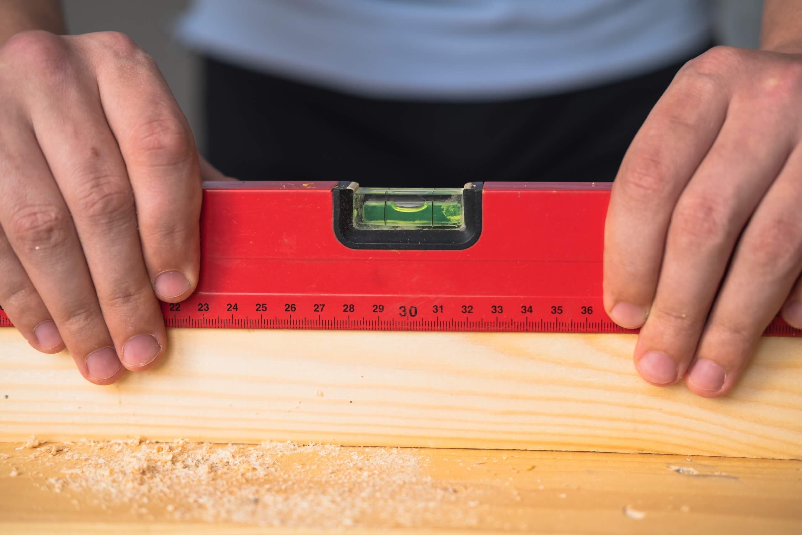 Two hands hold a red spirit level against a piece of wood. The spirit level, essential for precise TV wall mounting, also functions as a measuring tool with centimeter markings. The wood surface has some sawdust scattered around. The person’s torso is visible, but their face is not in the frame.