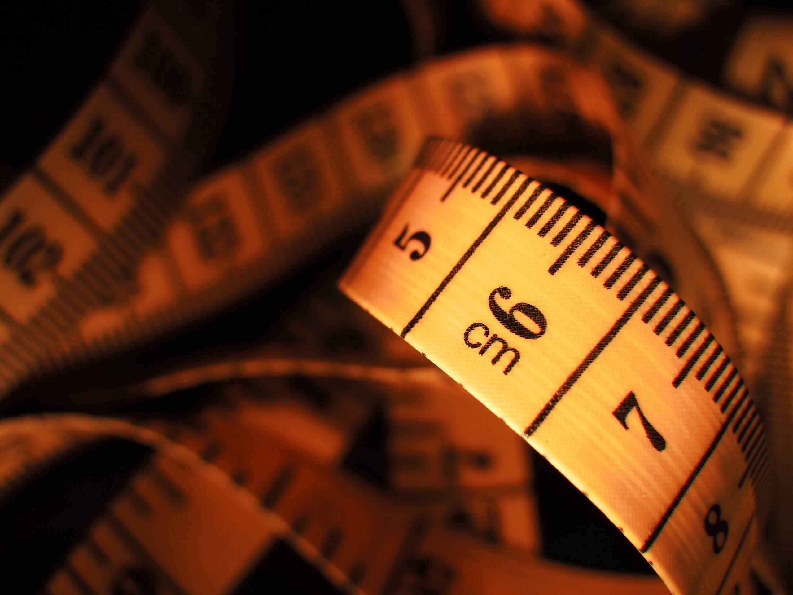 Close-up image of a coiled measuring tape, primarily showing the centimeter scale. The tape is illuminated with a warm light, highlighting the numbers and measurement markings clearly. The background is dark, creating high contrast with the tape—ideal for precise TV wall mounting measurements.