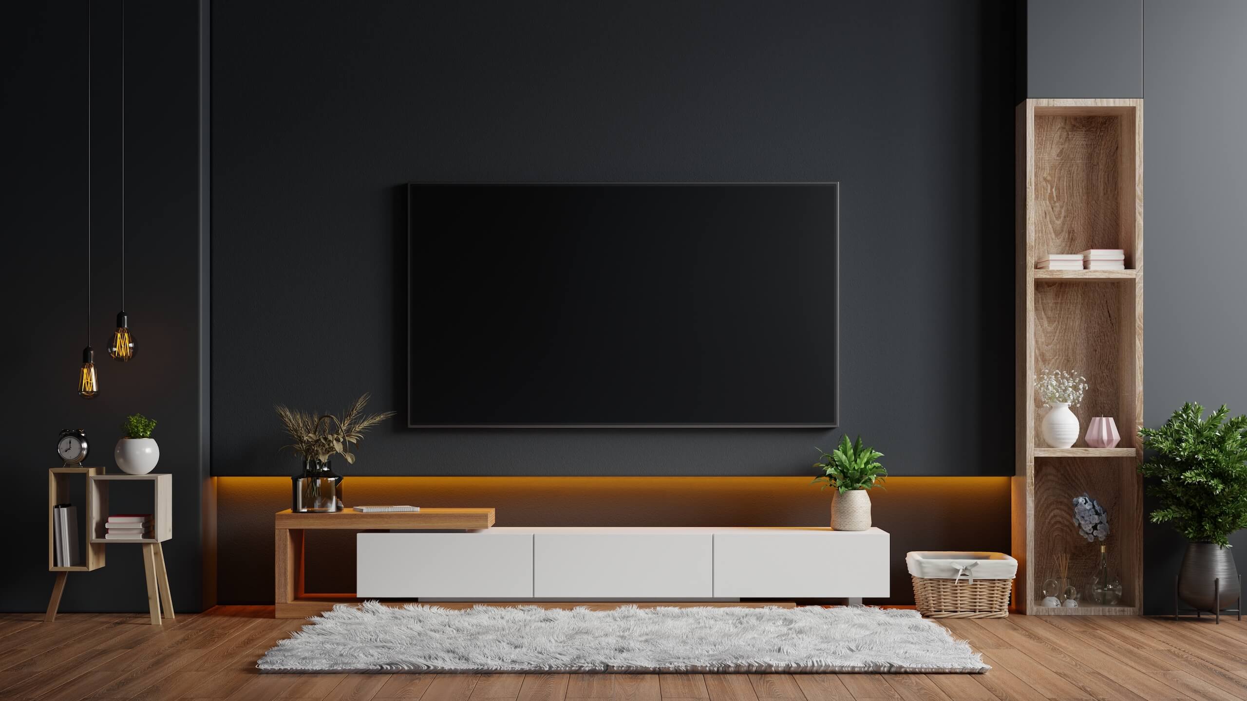 A modern, minimalist living room features TV Wall Mounting with a large flat-screen TV on a dark wall. Below, a white stand holds plants and decor. On the left, a small wooden side table boasts a lamp and clock, while to the right stands a tall wooden shelf adorned with plants and vases. A fluffy white rug covers the wooden floor.