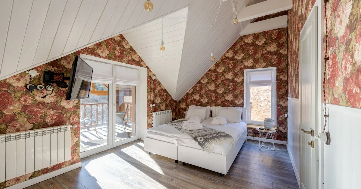 A cozy attic bedroom features floral-patterned wallpaper, a white slanted ceiling with hanging lights, a large window, and a double bed with white linens. A wall-mounted TV is positioned opposite the bed, and a balcony door provides natural light and outdoor access.