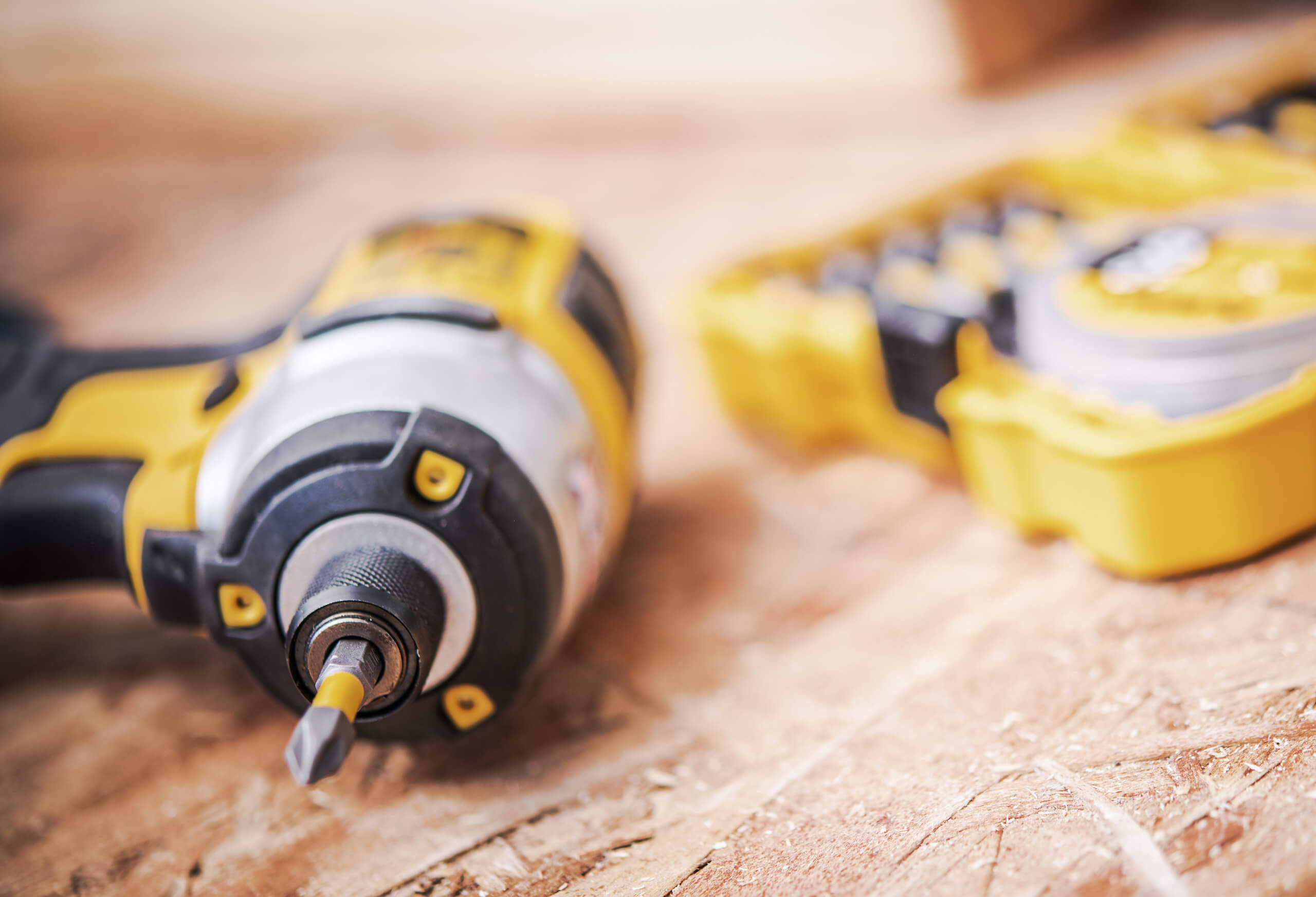 A close-up of a cordless power drill with a bit attached, placed on a wooden surface. In the blurry background, there is a yellow and black tool case containing various drill bits. The focus is on the drill, showcasing its details—perfect for tasks like TV wall mounting.