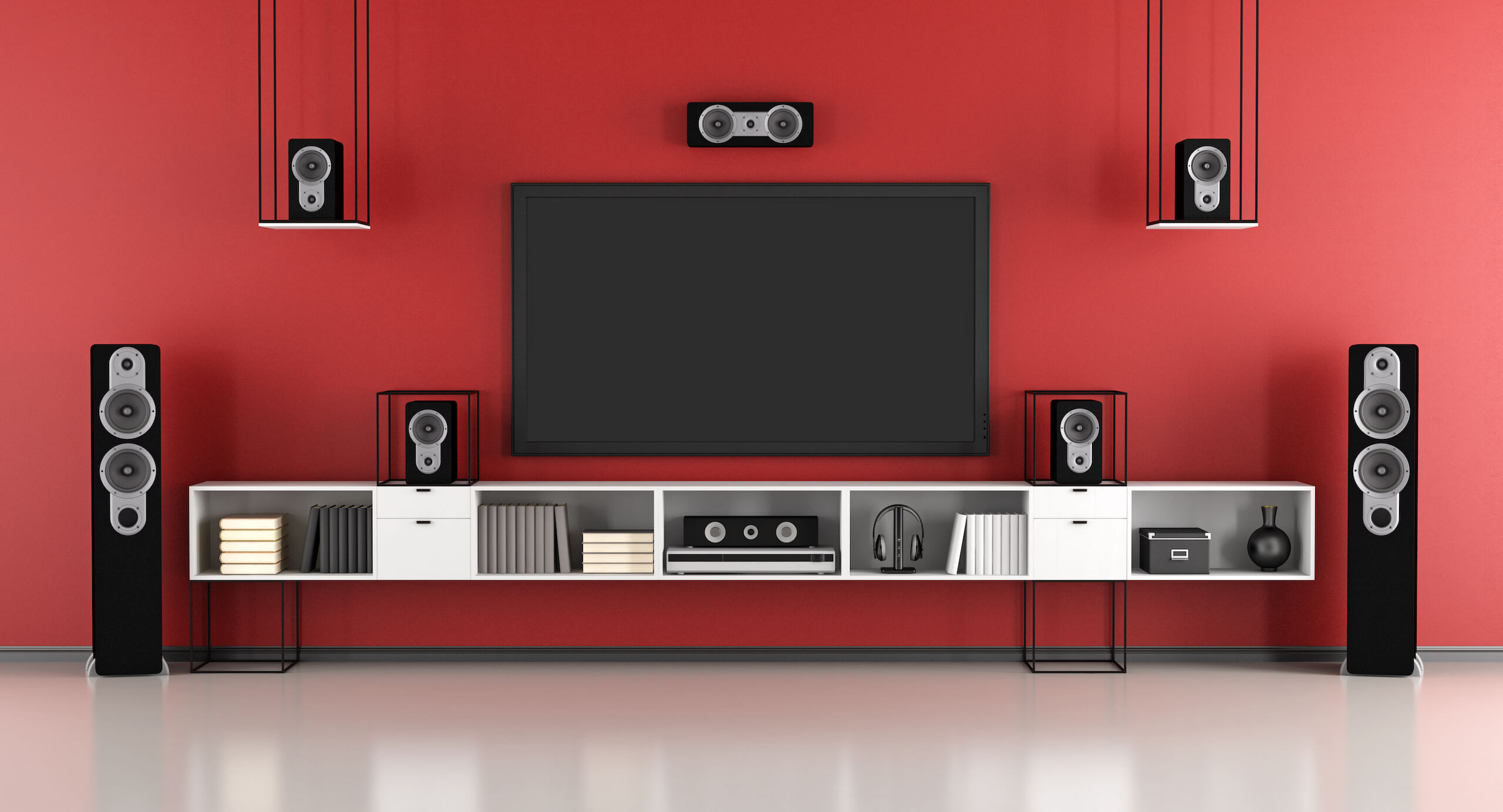 A modern home entertainment setup with a large flat-screen TV wall mounting on a red wall. Surrounding the TV, there are five black speakers: two tall floor-standing speakers, two smaller ones on shelves, and one center speaker above the TV. The shelves house books and decorative items.