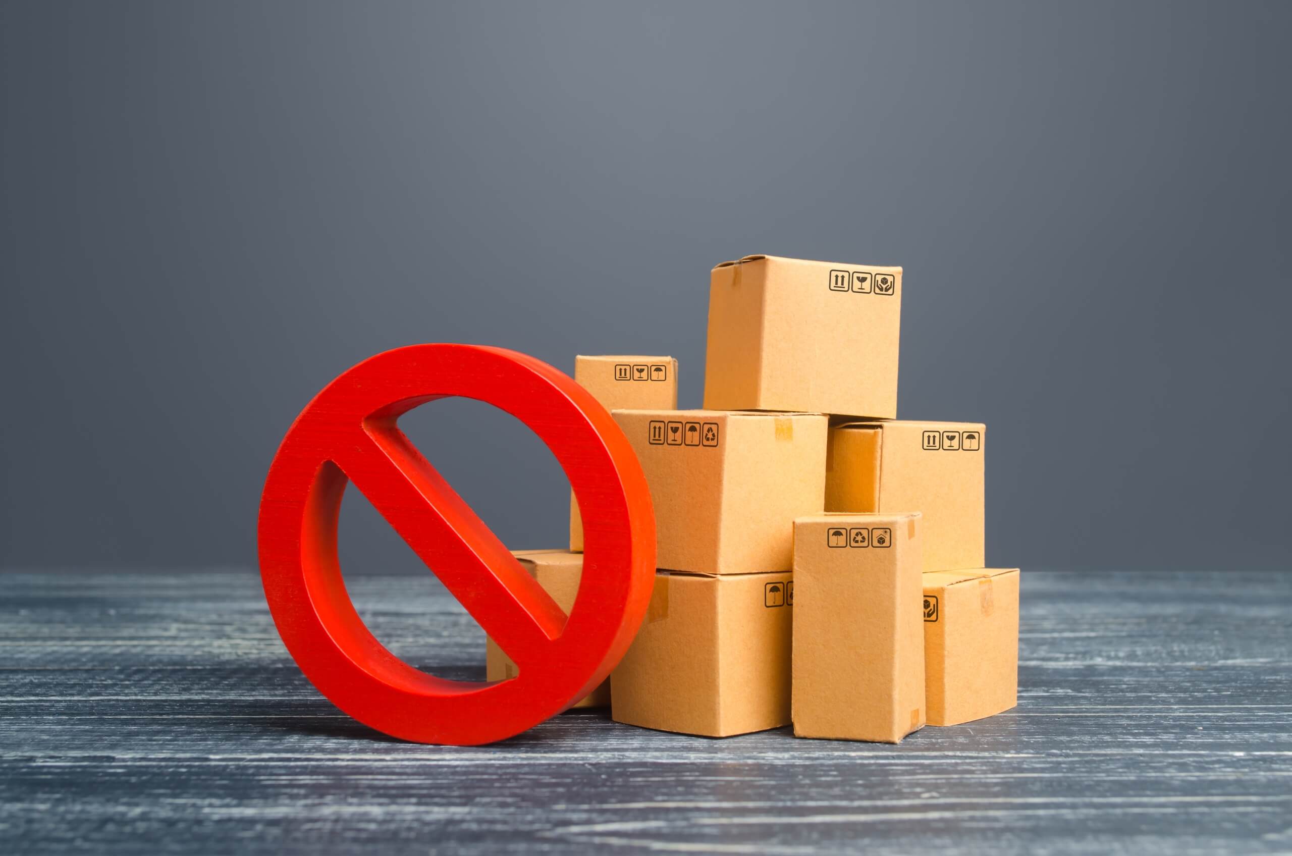 A group of small cardboard boxes is stacked together on a wooden surface. To the left of the boxes, there's a red circle with a diagonal line through it, resembling a "No" or "Prohibited" symbol. The plain gray wall in the background suggests an unadorned area perfect for TV wall mounting.