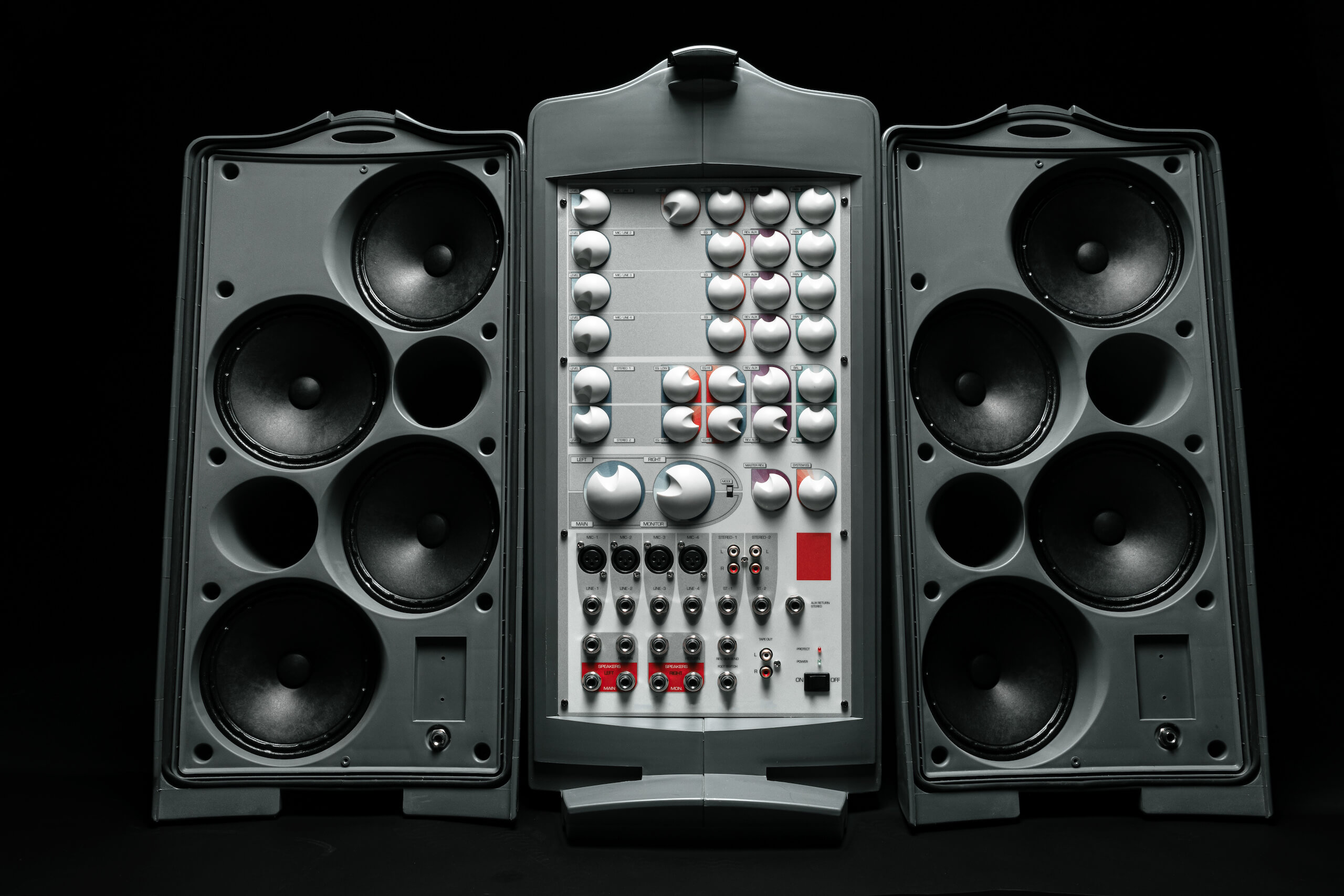 A detailed photo of an audio equipment setup featuring two large black speaker units with several circular components on both sides. In the center, there is a control panel with various buttons, knobs, and switches. Nearby, a TV wall mounting bracket subtly complements the sleek background.