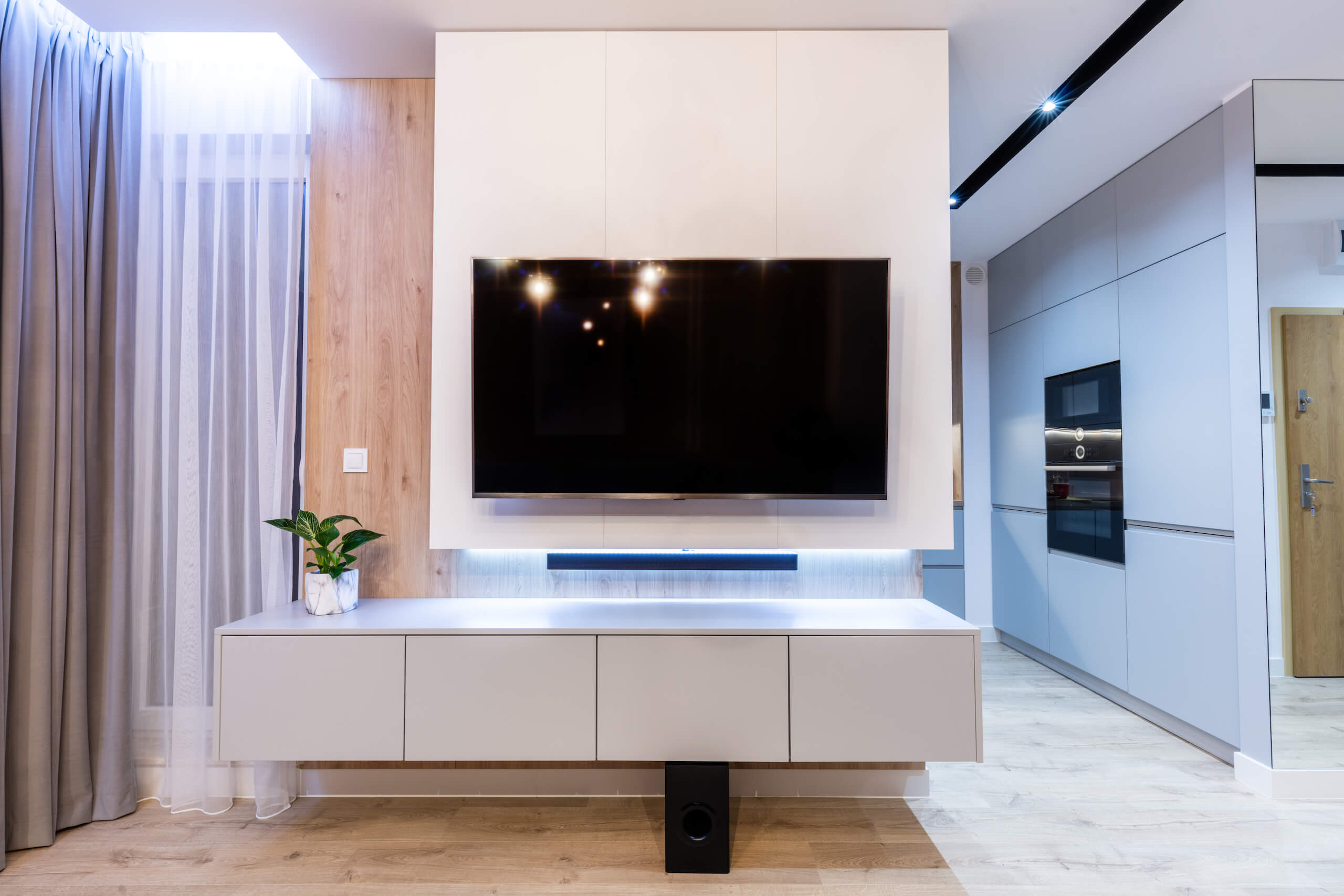 A modern living room showcases a large flat-screen TV with expert TV wall mounting above a sleek, white media console. A soundbar is placed below the TV, with a small plant on the left side of the console. The space is bright, featuring light wood accents and floor.