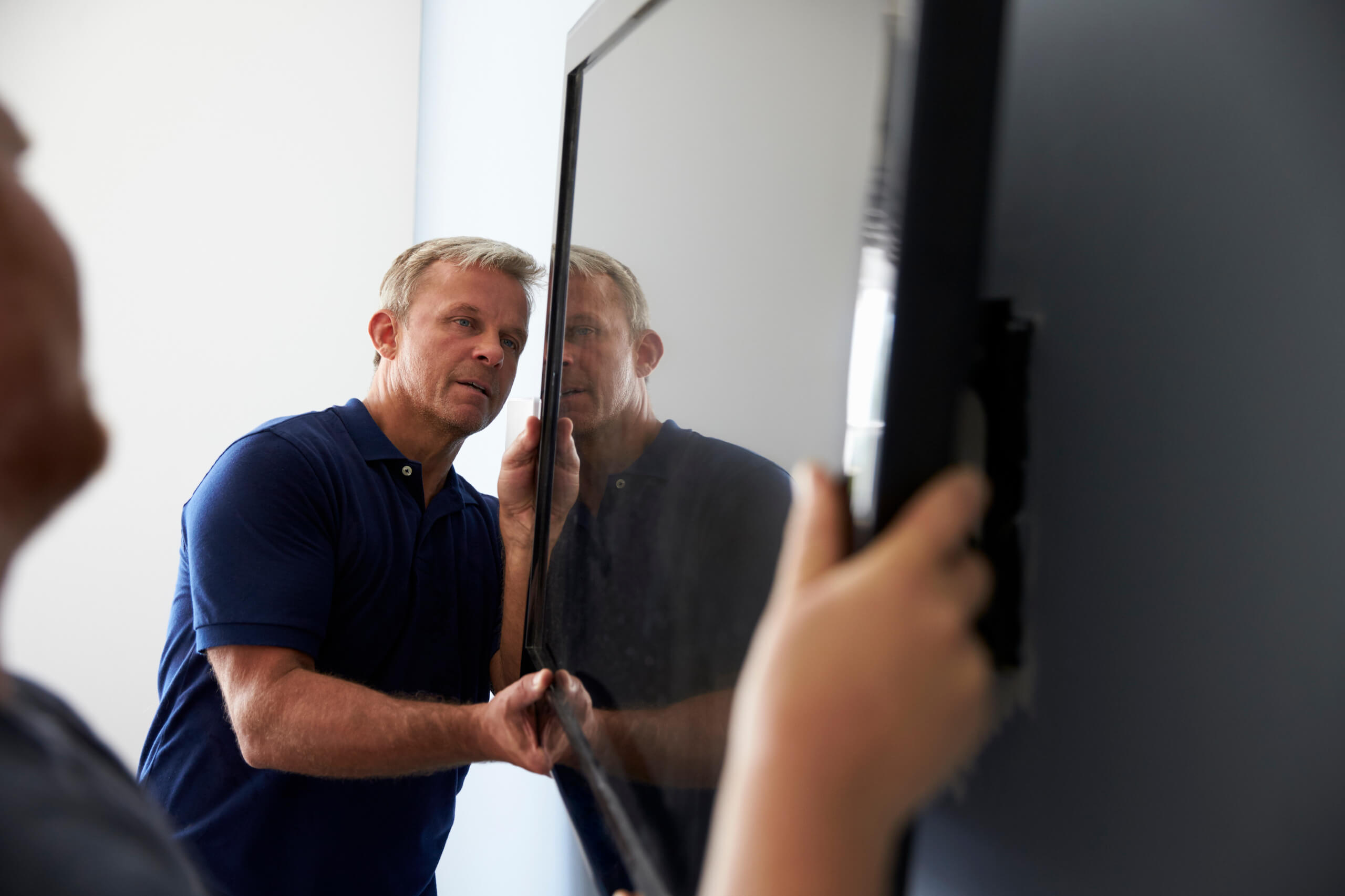 Two men are installing a flat-screen TV on a wall. One man appears to be securing the TV wall mounting while the other steadies the screen, both focused on the task. They are in a well-lit room with a plain white and grey background.