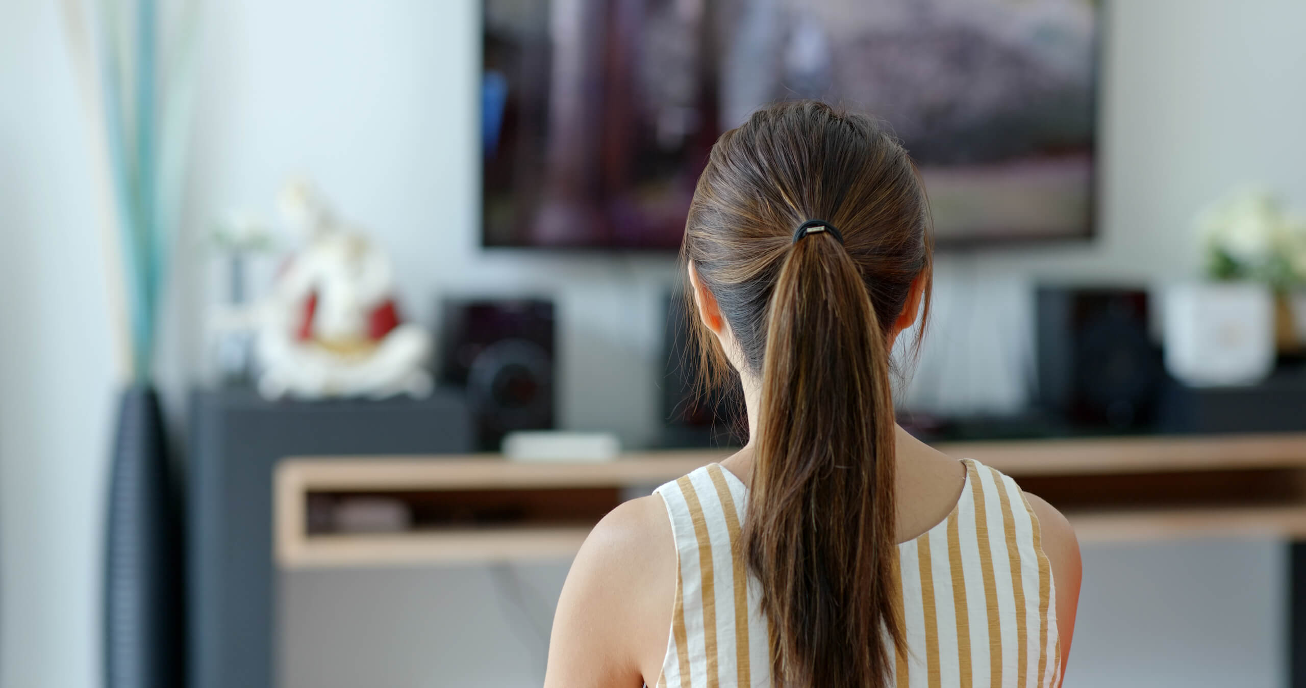 A person with long brown hair tied in a ponytail is sitting indoors and facing a television screen mounted on the wall. The background includes a blurred shelf with various items, and the person is wearing a sleeveless white and yellow striped top.